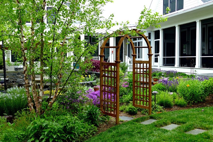 Lush garden with colorful flowers, a wooden arbor, and a path leading to a white house with a large porch.